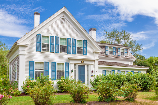 New England House with White Clapboard Exterior and Grey Shingle Roof, Blue Window Shutters and Landscaped Front Yard with green grass, bushes and Flowers. The house is in Chatham, Cape Cod, Massachusetts. Canon EF 24-105mm/4L IS USM Lens.