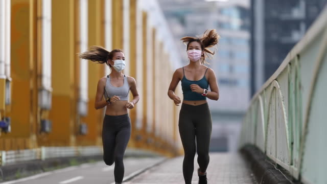 Two women wearing masks jogging on city bridge with buildings background in the morning, slow motion