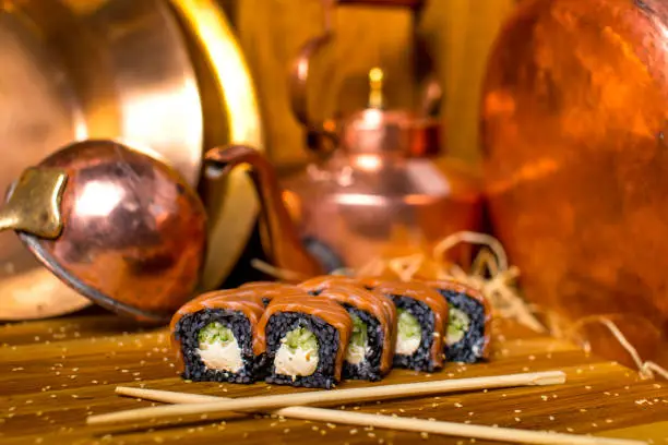 Japanese Philadelphia sushi roll with black rice, raw salmon fillet on wooden table served with sesame seeds with copper tableware on background. Chopsticks near pan Asian seafood dish. Still life