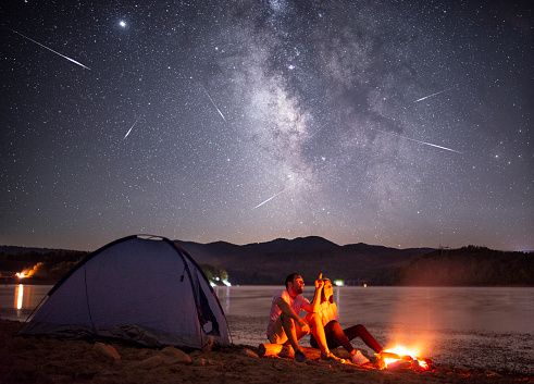 Side view of loving couple sitting near bright burning campfire and tent, enjoying beautiful camping night together under dark sky full of shiny stars and bright Milky Way, warm summer night. They watching the Perseid meteor shower