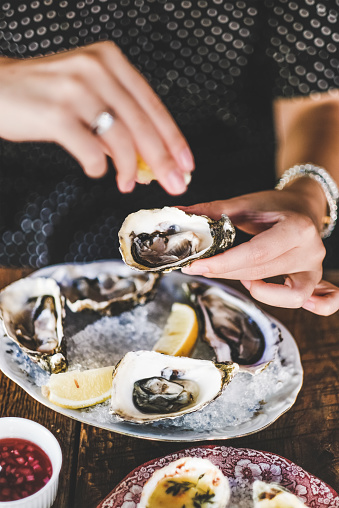 Hands of woman squeezing lemon juice to fresh Irish oysters over ice in plate in fish restaurant, selective focus. Seafood, French cuisine, fine dining concept
