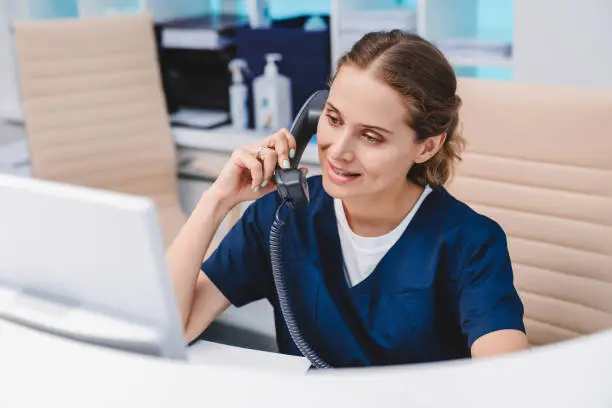 Photo of Young female receptionist talking on phone in clinic while sitting and looking on pc monitor