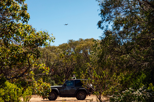A shot of a mid adult male standing out the open top of a 4x4 vehicle in a rural, remote location. He is wearing casual clothing and flying a drone.
