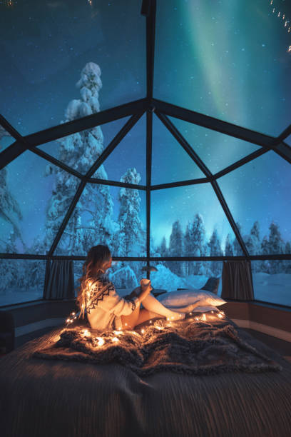 Young woman enjoying a view of the northern lights Young traveler woman standing on the comfortable bed and watching breathtaking northern lights or Aurora Borealis over a snowy landscape in starry night in Lapland, Finland finland stock pictures, royalty-free photos & images