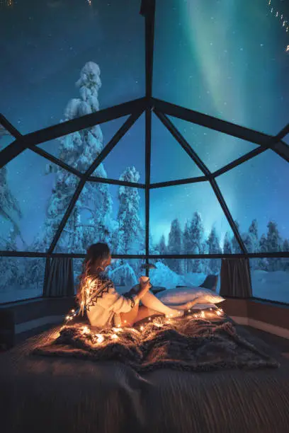 Young traveler woman standing on the comfortable bed and watching breathtaking northern lights or Aurora Borealis over a snowy landscape in starry night in Lapland, Finland