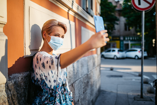 Young Caucasian beautiful woman with protective face mask making selfie on the sidewalk.