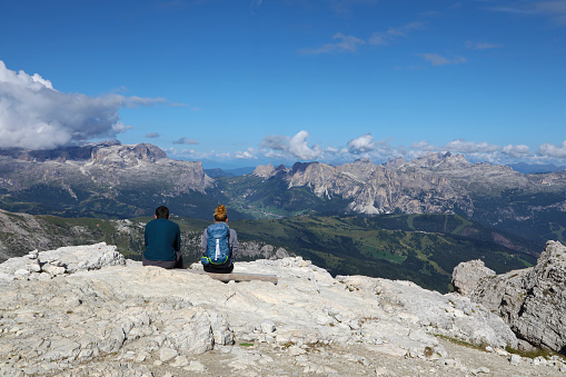 Two seated young people look at a breathless landscape in front of them from the top of Mount Lagazuoi in italian Alps.