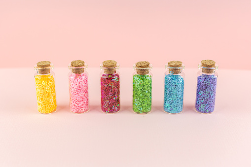 Small glass bottles or jars with sealed corks with multicolored shiny stars stand in a row on a pink background. Sequins decoration for creativity, needlework. Storage and order concept.