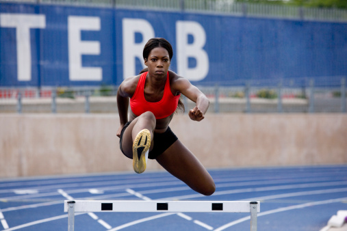 Female fit caucasian runner in sportswear running fast and jumping through hurdle at stadium track wide shot