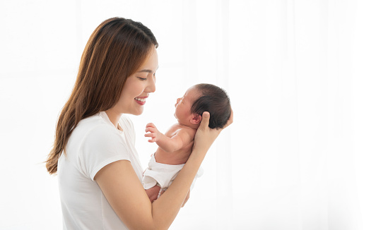 Pretty asian woman holding a newborn baby in her arms. Happy family. Laughing mother lifting her adorable infant baby son on white.