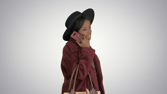 Medium shot. Side view. African american fashion girl in coat and black hat talking on the phone while walking on gradient background. Professional shot in 4K resolution. 046. You can use it e.g. in your medical, commercial video, business, presentation, broadcast