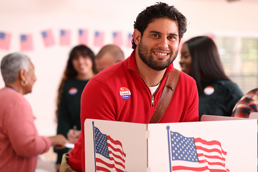 Mid-adult, Latin descent man votes in the USA election.  He stands at voting booth in polling station.   Other voters and election day registration seen in background.