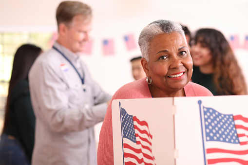 African descent, senior adult woman votes in the USA election.  She stands at voting booth in polling station.   Other voters and election day registration seen in background.