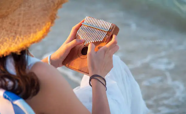 Lonely female playing kalimba music instrument by the seaside while sitting on the beach tanning chair at sunset and enjoying free time time alone