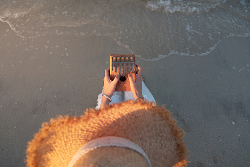 Lonely female playing kalimba music instrument by the seaside while sitting on the beach tanning chair at sunset and enjoying free time time alone top high angle view
