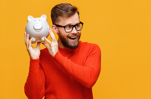 Optimistic bearded man in glasses looking away with opened mouth and shaking piggy bank with money near ear against yellow background