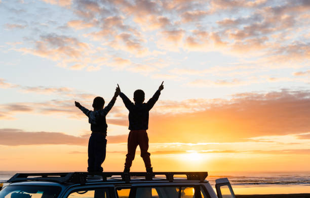 Kids with arms outstretched on top of 4wd rood. Kids at beach standing on the roof of 4wd and enjoying sunset. rood stock pictures, royalty-free photos & images
