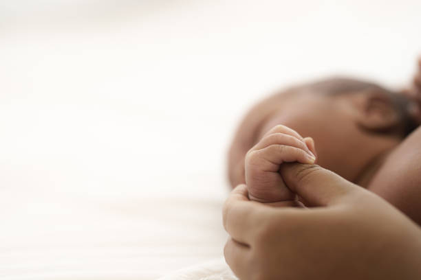 母の手が赤ちゃんを引き上げる間、ベッドに横たわっているアフリカ系アメリカ人の幼児の赤ちゃん - 誕生 ストックフォトと画像