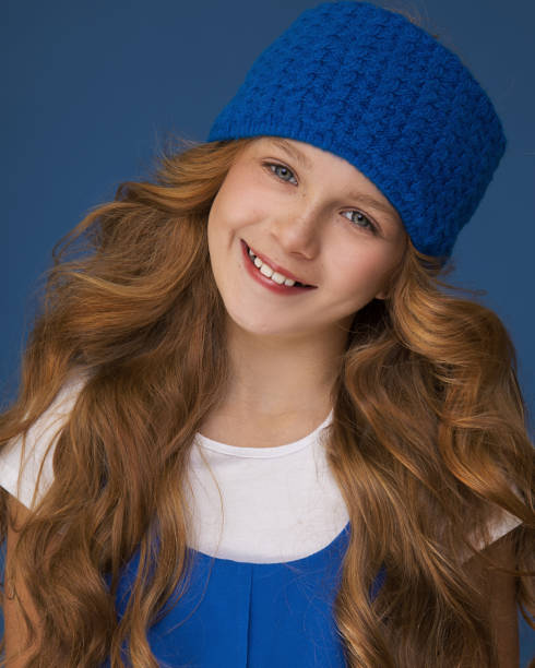 カールとそばかすに豪華な赤い髪を持つ10歳の若い十代の少女のクローズアップ肖像画。青い背景でスタジオのカメラを笑顔で見ます。暖かい帽子のアクセサリーの概念。 - 10 11 years child human face female ストックフォトと画像