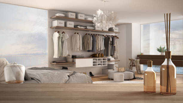 istock Wooden table top or shelf with aromatic sticks bottles over blurred modern bedroom with double bed, carpet, walk-in closet and panoramic window, white architecture interior design 1275772502