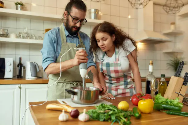 Italian man, chef cook using hand blender while preparing a meal. Young woman, girlfriend in apron looking at the process, helping in the kitchen. Cooking at home, Italian cuisine. Horizontal shot