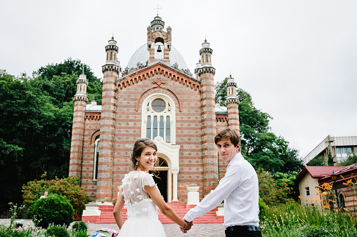 Bride dressed in white dress and groom in suit going together. Newlyweds going to church before wedding ceremony.