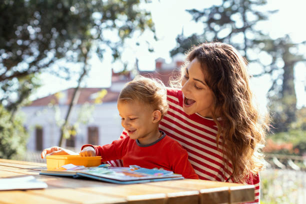 mãe feliz e criança lendo livro infantil lift-the-flap e comendo lanches no jardim - looking at camera smiling desk isolated - fotografias e filmes do acervo