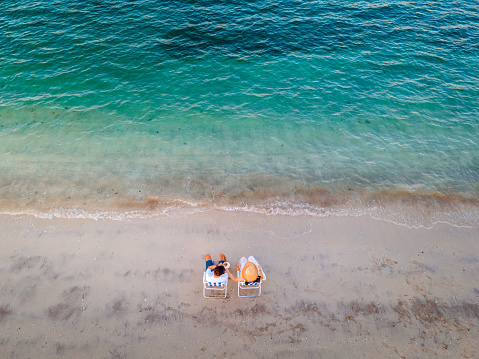 Couple sharing coconut water and relaxing on the beach sitting on the tanning chairs at sunset top down aerial view with copy space