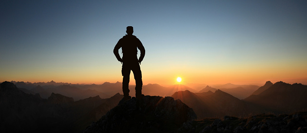 Happy winning successful man silhouette at sunset or sunrise standing relaxed and is happy for having reached mountain peak summit goal during hiking travel trek. Tirol, Austria and Allgäu Alps, Bavaria, Germany