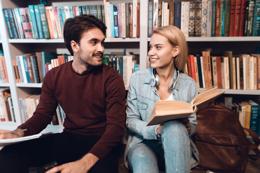 Young girl with a book sits in a library. A man and a girl are sitting on the floor in the library and looking at each other.