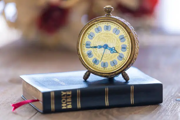 Old alarm clock on Holy Bible with flowers on wooden table. Focus on clock