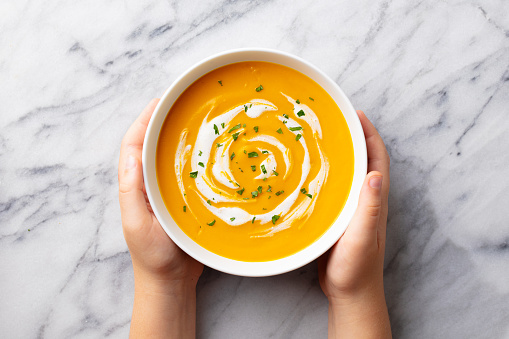 Kids hands holding a bowl with pumpkin soup. Marble background. Top view.