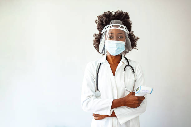 Confident middle aged woman doctor in safety face mask and white coat holding infrared thermometer, standing isolated on white background. Epidemic preventionPublic healthMeasuring body temperaturescreening
