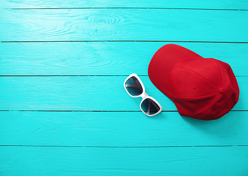 Red cap and sunglasses on blue wooden background. Top view and copy space. Summertime accessories.