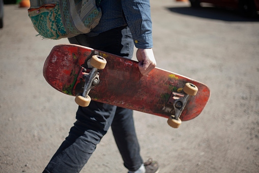 Skateboard in the guy's hand. The man carries the board in his hand. The guy on a sunny day goes to the skateboard.