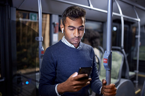 Indian young business man with earphones working using smartphone on public bus