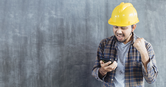 Construction worker man holding smartphone and celebrating his success over gray background with copy space