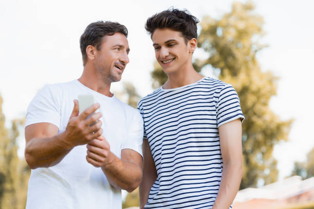 joyful teenager looking at smartphone in hands of father - teenager parent father son imagens e fotografias de stock
