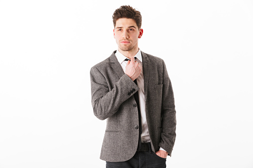 Calm business man in jacket with arm in pocket corrects his tie and looking at the camera over white background