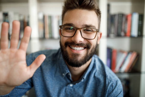 Smiling young man having online conference from home office Smiling young man having online conference from home office waving gesture stock pictures, royalty-free photos & images
