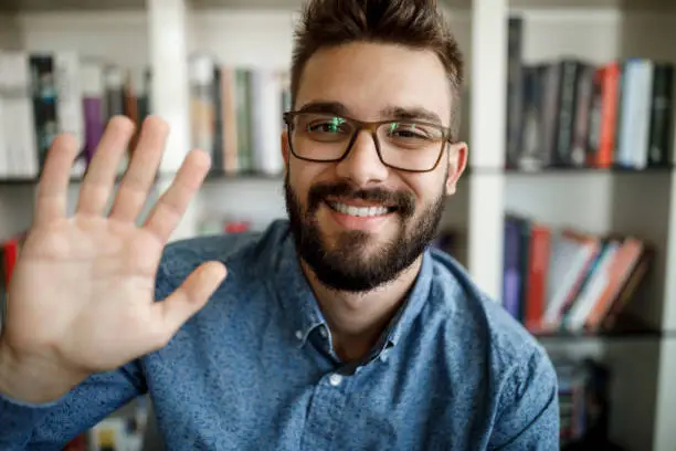 Photo of Young man waving with hand on video call at home office