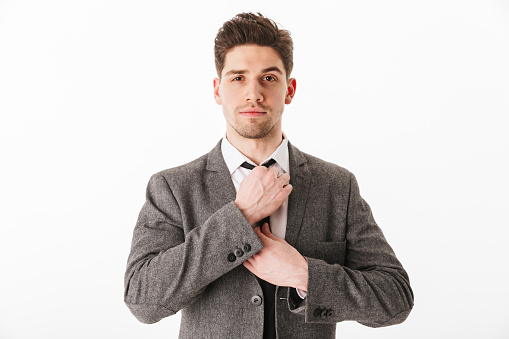 Calm business man in jacket corrects his tie and looking at the camera over white background