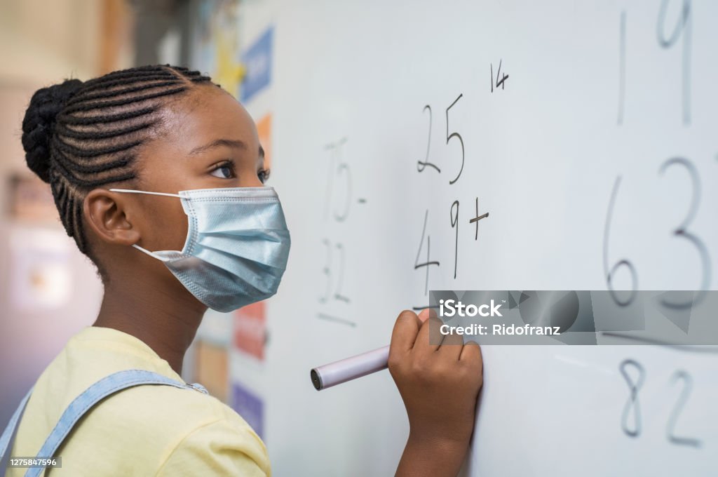 Elementary girl wearing protective face mask at school Portrait of african girl wearing face mask and writing solution of sums on white board at school. Black schoolgirl solving addition sum on white board during Covid-19 pandemic. School child thinking while doing mathematics problem and wearing surgical mask due to coronavirus. Child Stock Photo