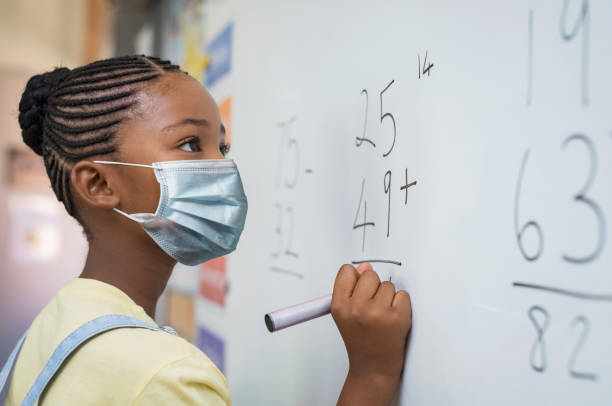 Elementary girl wearing protective face mask at school Portrait of african girl wearing face mask and writing solution of sums on white board at school. Black schoolgirl solving addition sum on white board during Covid-19 pandemic. School child thinking while doing mathematics problem and wearing surgical mask due to coronavirus. back to school photos stock pictures, royalty-free photos & images