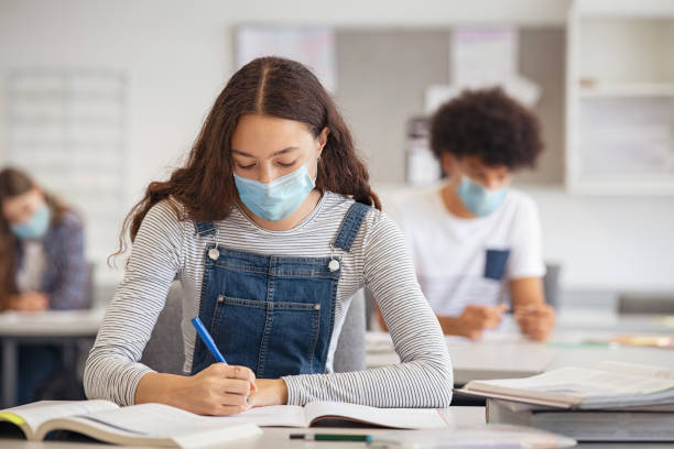 High school girl studying in class with face mask High school student taking notes from book while wearing face mask due to coronavirus emergency. Young woman sitting in class with their classmates and wearing surgical mask due to Covid-19 pandemic. Focused girl studying in classroom completing assignment during corona virus. homework photos stock pictures, royalty-free photos & images