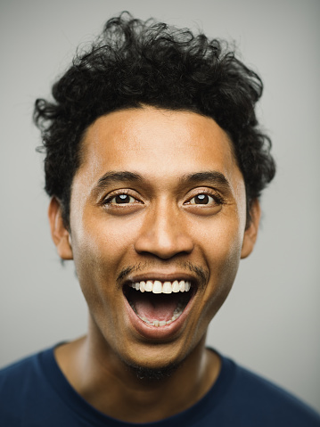 Close up portrait of young adult middle eastern man with shouting expression against white gray background. Vertical shot of real pakistani man smiling in studio with short black hair. Photography from a DSLR camera. Sharp focus on eyes.