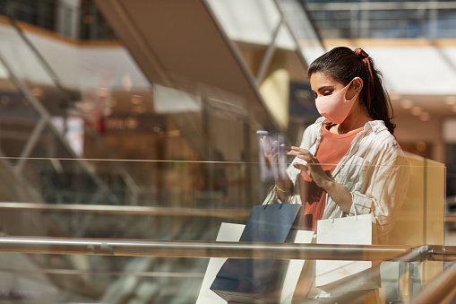 Side view portrait of young woman wearing mask while shopping in mall and using smartphone, copy space