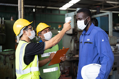 Group of factory workers wearing protective face mask and using infrared forehead thermometer before start of work in front of the industrial factory