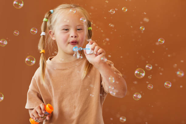 Girl with Down Syndrome Blowing Bubbles Waist up portrait of cute girl with down syndrome blowing bubbles while posing against brown background in studio, copy space down syndrome stock pictures, royalty-free photos & images