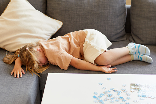 Full length portrait of cute girl with down syndrome falling asleep on sofa while playing board games at home, copy space